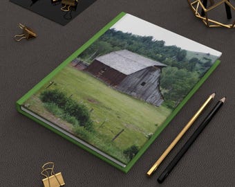 Könnte beinhalten: Ein grünes Journal mit einem Foto einer Holzscheune auf einem Feld mit grünen Bäumen und Gras. Das Journal liegt auf einer braunen Oberfläche mit goldenen Büroklammern und Bleistiften.