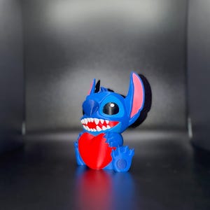 K&ouml;nnte beinhalten: Blaue Stitch-Figur, die ein rotes Herz h&auml;lt. Die Figur hat gro&szlig;e Ohren mit rosa Innendetails, ein breites Grinsen mit wei&szlig;en Z&auml;hnen und schwarze Augen. Die Figur stammt aus Lilo & Stitch. Auf dunkler Oberfl&auml;che.