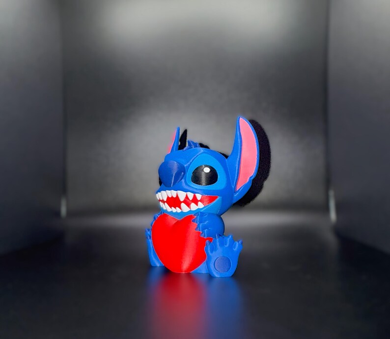 K&ouml;nnte beinhalten: Blaue Stitch-Figur, die ein rotes Herz h&auml;lt. Die Figur hat gro&szlig;e Ohren mit rosa Innendetails, ein breites Grinsen mit wei&szlig;en Z&auml;hnen und schwarze Augen. Die Figur stammt aus Lilo & Stitch. Auf dunkler Oberfl&auml;che.