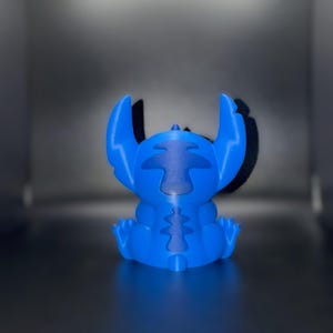 K&ouml;nnte beinhalten: Blaue Figur der Figur Stitch aus dem Zeichentrickfilm Lilo & Stitch. Die Figur sitzt mit ausgestreckten Armen und Beinen. Die Ohren sind gro&szlig; und spitz. Die Figur besteht aus einem einfarbigen blauen Material.
