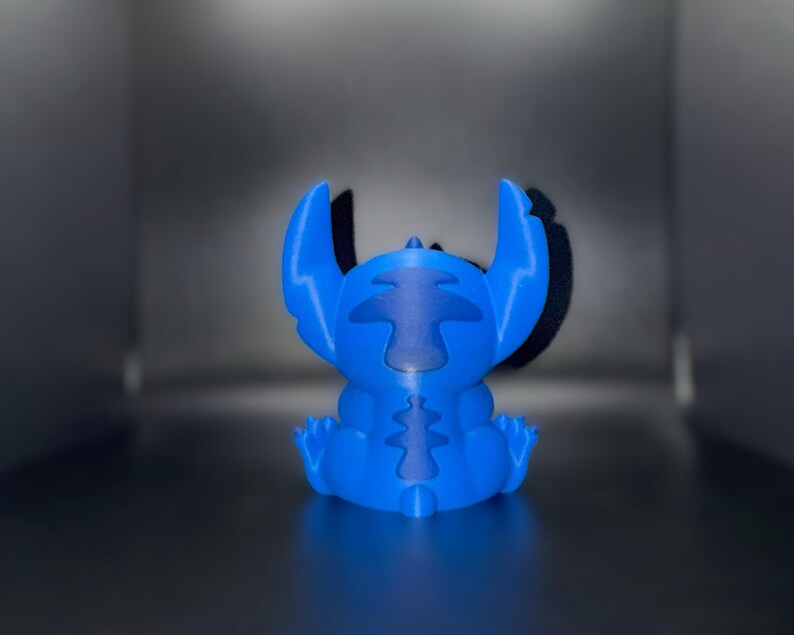 K&ouml;nnte beinhalten: Blaue Figur der Figur Stitch aus dem Zeichentrickfilm Lilo & Stitch. Die Figur sitzt mit ausgestreckten Armen und Beinen. Die Ohren sind gro&szlig; und spitz. Die Figur besteht aus einem einfarbigen blauen Material.