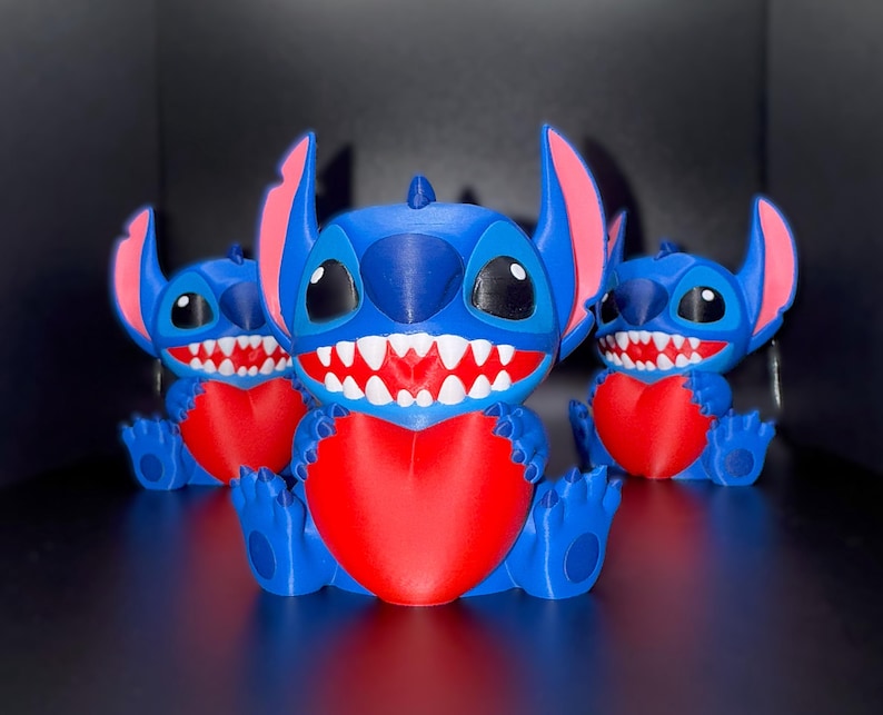 K&ouml;nnte beinhalten: Drei blaue und rote Stitch-Figuren sind in einer Reihe angeordnet. Jede Figur hat gro&szlig;e Ohren, ein breites Grinsen und h&auml;lt ein rotes Herz. Die Figuren sitzen auf einer reflektierenden Oberfl&auml;che.