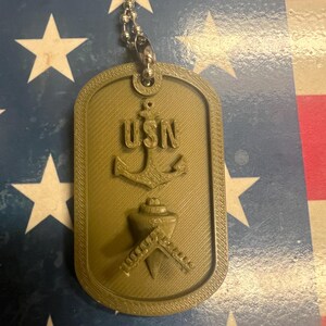 May include: A green metal dog tag with the letters USN, an anchor, and a crossed compass and plumb bob. The dog tag is on a silver chain.