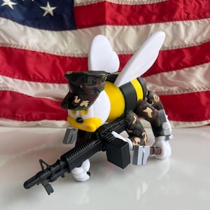 May include: A novelty figurine of a bee-like character in a camouflage military uniform and hat, holding a black rifle. The bee has a yellow body with black stripes, white wings, and a duck-like face. The background is an American flag.