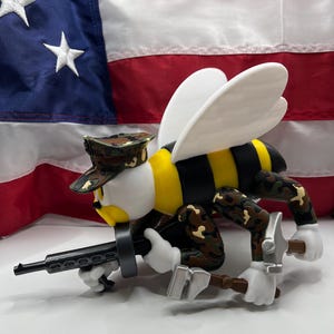 May include: A whimsical figurine of a bee in a camouflage military uniform, holding a machine gun and a hammer. The bee has white wings, a yellow and black striped body, and a camouflage hat. The background features an American flag.