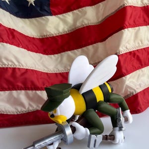 May include: A figurine of a bee-like character in a military uniform, holding a silver weapon. The figure has a yellow and black striped body, white wings, and a green cap. The background features an American flag.