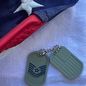 May include: Two green military dog tags on a silver chain. One tag has a blue star and stripes, the other has the American flag.