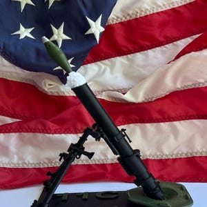 May include: A black mortar model with a green projectile, set against an American flag. The base of the mortar has the text "WE Bring Our Own Rain". The flag features white stars on a blue field and red and white stripes.