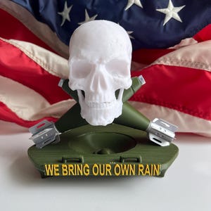 May include: A white skull figurine rests on a green base with two green missile-shaped accents. The base has the text "WE BRING OUR OWN RAIN!" in yellow. An American flag serves as the backdrop.