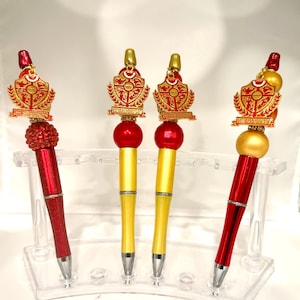 May include: Four pens featuring red and gold metallic barrels, topped with large spherical beads and a gold-toned crest emblem. The crest displays intricate symbols and the text "NATIONAL SORORITY OF PHI DELTA KAPPA INC" in red and gold.