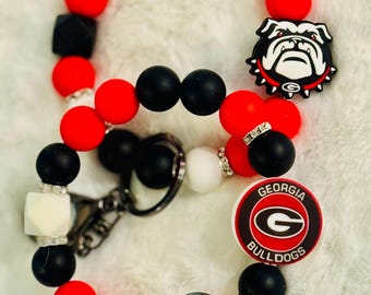 May include: A beaded keychain and wristlet set in red, black, white, and silver. The set features a Georgia Bulldogs logo and bulldog charm. The beads are round and square, with silver rhinestone accents.