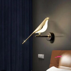 May include: A gold bird-shaped wall sconce with a black metal base. The sconce is mounted on a wall and is illuminating a nearby bed.