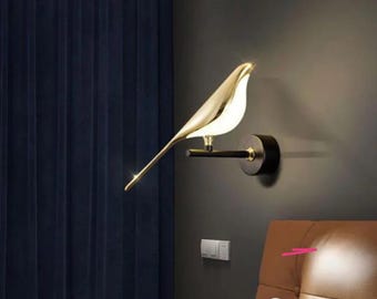 May include: A gold bird-shaped wall sconce with a black metal base. The sconce is mounted on a wall and is illuminating a nearby bed.