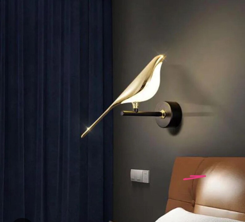 May include: A gold bird-shaped wall sconce with a black metal base. The sconce is mounted on a wall and is illuminating a nearby bed.