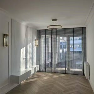May include: A modern living room with white walls, a white ceiling, and a white built-in shelf. The room has a large window with black blinds and a view of a city. The floor is made of wood planks in a herringbone pattern.