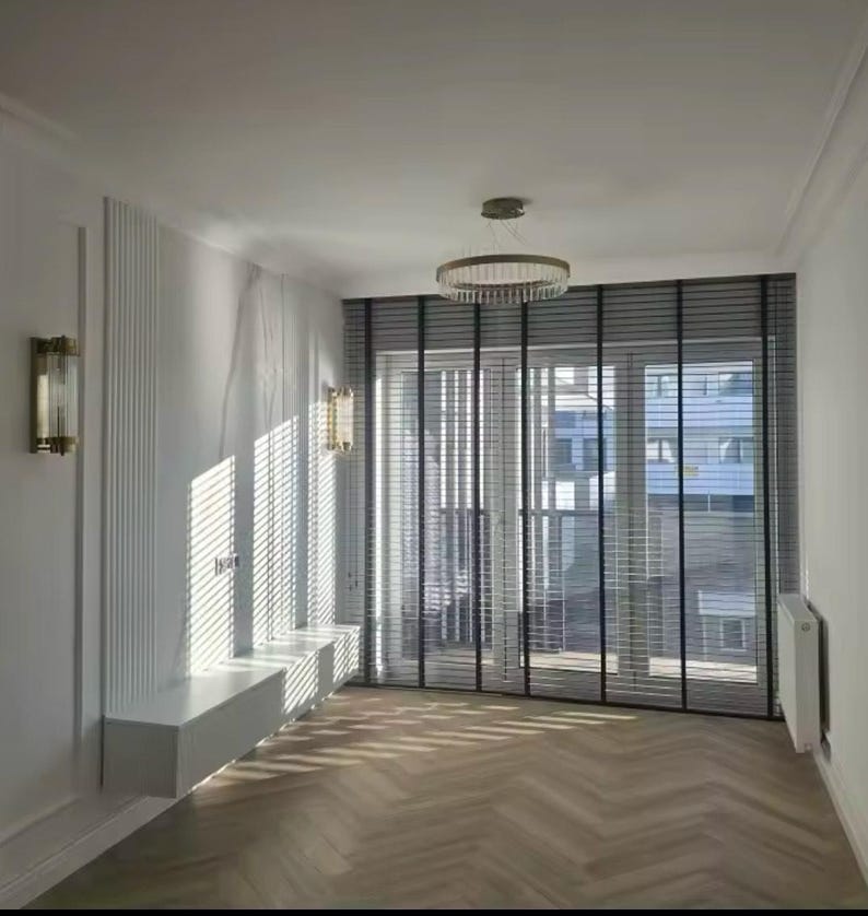 May include: A modern living room with white walls, a white ceiling, and a white built-in shelf. The room has a large window with black blinds and a view of a city. The floor is made of wood planks in a herringbone pattern.