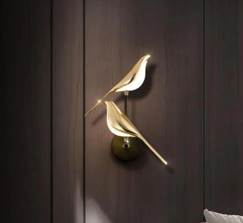 May include: A pair of gold bird-shaped wall sconces with a modern design. The sconces are mounted on a wall and have a minimalist aesthetic.