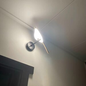 May include: A white bird-shaped wall sconce with a black metal base and a white light bulb. The sconce is mounted on a white wall.