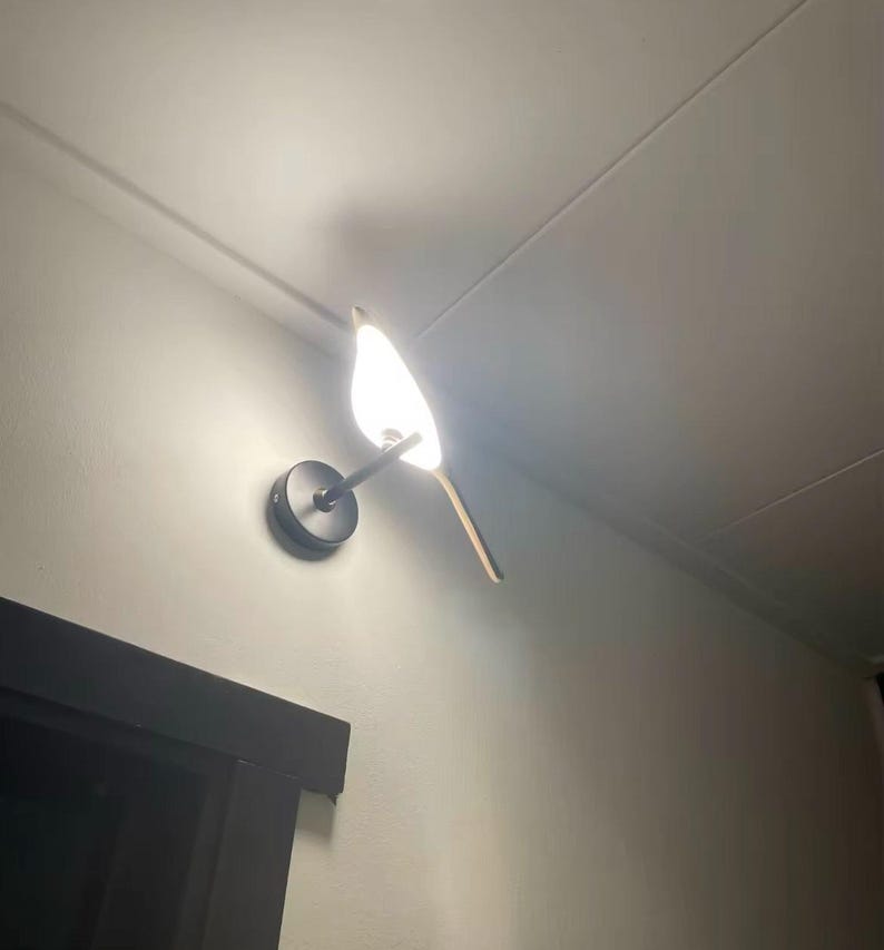 May include: A white bird-shaped wall sconce with a black metal base and a white light bulb. The sconce is mounted on a white wall.