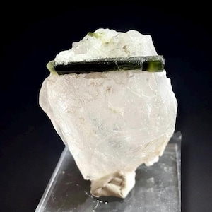 May include: A clear quartz crystal with a dark green tourmaline crystal on top. The quartz is mounted on a clear acrylic display stand. The tourmaline is a dark green color with a rectangular shape. The quartz crystal is a light, opaque color.