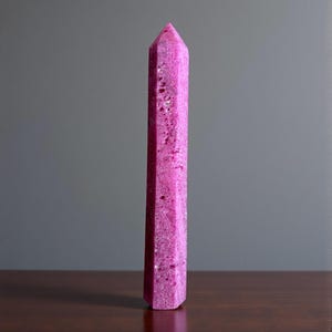 May include: A tall, pink crystal obelisk with a pointed top. The crystal has a textured surface and is standing on a dark wooden surface. The obelisk is a vibrant shade of pink and is set against a gray background.