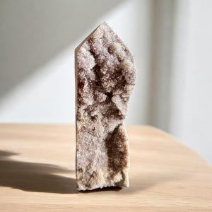 Puede incluir: Una geoda de cristal de amatista natural de pie, con una superficie rugosa y texturizada. El cristal es una mezcla de tonos morados claros y beige, con una estructura cristalina. La geoda se exhibe sobre una superficie de madera clara.