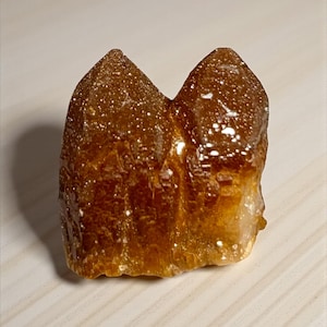 May include: A raw, natural citrine crystal cluster. The crystal has a warm, amber color with a sparkling, textured surface. The shape is irregular, with two prominent peaks. The crystal sits on a light-colored surface.
