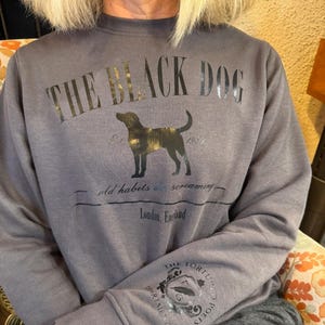 Peut inclure: Un sweat-shirt gris avec le texte "THE BLACK DOG" imprimé sur le devant. Le texte est en noir et la silhouette du chien est en or. Le texte "Est. 1959" est imprimé sous le chien. Le texte "old habits die screaming" est imprimé sous la date. Le texte "London, England" est imprimé sous la citation. Le texte "THE FORTUNE OF POETS" est imprimé sur la manche gauche.