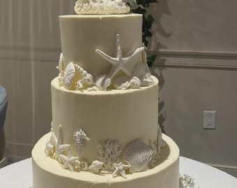 May include: A three-tiered wedding cake with ivory frosting, decorated with white seashells, starfish, seahorses, and coral. The top tier features a seahorse cake topper. The cake sits on a table with floral accents, creating a beach-themed design.