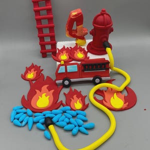 May include: A festive cake decoration set featuring a red fire truck, a red fire hydrant, and a yellow hose. The set includes red flames, blue water droplets, a red ladder, and a number 4. The decorations are likely made of fondant or gum paste.