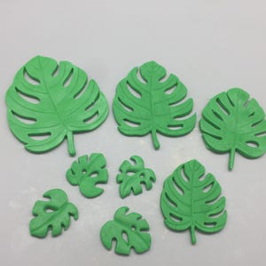 May include: A collection of eight green, artificial Monstera leaves in various sizes. The leaves have a detailed, veined texture and are arranged on a white surface. The leaves are a vibrant shade of green.
