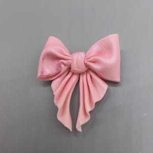 May include: A light pink bow with a velvety texture. The bow has a classic shape with two loops and two long, flowing tails. The bow is centered and appears to be made of a soft, pliable material.
