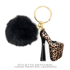 Könnte beinhalten: Ein Stiletto-Schlüsselanhänger mit goldenem Ring und einem schwarzen, flauschigen Bommel. Der High Heel ist handbemalt mit einem Leopardenmuster. Der Text unten lautet "Stiletto Keychain Hand Painted Leopard".