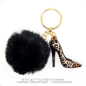 Può includere: Un portachiavi a stiletto con un pompon nero e un portachiavi color oro. Il tacco alto è dipinto a mano con un motivo leopardato. Il testo in basso recita: "Stiletto Keychain Hand Painted - Leopard."
