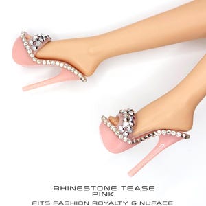 May include: A pair of light pink high-heeled sandals featuring a clear strap adorned with sparkling clear rhinestones along the edges and across the top. The text below reads RHINESTONE TEASE PINK FITS FASHION ROYALTY & NUFACE.