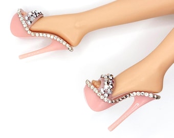 May include: A pair of light pink high-heeled sandals featuring a clear strap adorned with sparkling clear rhinestones along the edges and across the top. The text below reads RHINESTONE TEASE PINK FITS FASHION ROYALTY & NUFACE.
