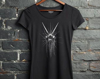May include: A black short-sleeved t-shirt featuring a white graphic illustration of a creature with multiple eyes and radiating lines. The shirt has a round neckline and is displayed on a wooden hanger. The text WRATH IN THE MACHINE is visible.