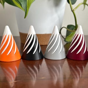 May include: Eight cone-shaped ring holders in different colours, including red, green, orange, black, grey, burgundy, purple, and lavender. Each cone has a white swirl design.