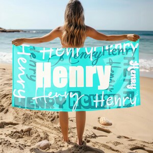 Person stands on a sandy beach holding a bright blue personalized beach towel with the name Henry in different fonts. Custom name towel, perfect for pool, swim, or summer trips, and a thoughtful gift for any age.