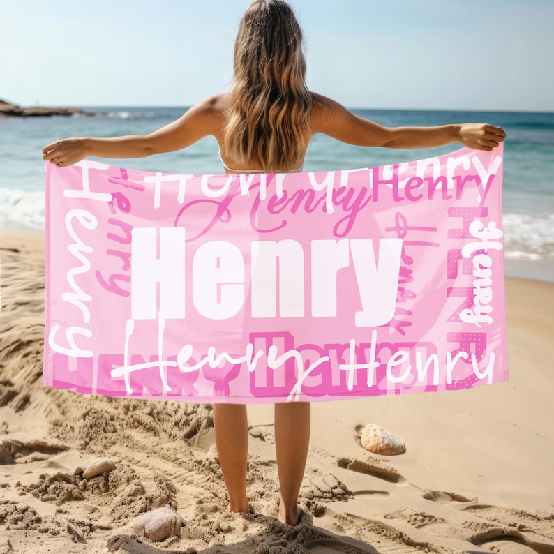Person stands on a sandy beach holding a pink personalized beach towel with the name Henry in large and small fonts. Custom name towel, perfect for pool, swim, or summer trips, and a thoughtful gift for all ages.