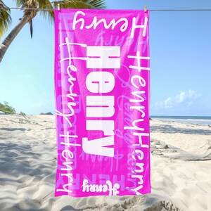 Bright pink personalized beach towel with the name Henry in different fonts, hanging on a line at the beach with palm trees and sand. Custom name towel, perfect for pool, swim, or summer trips for all ages.