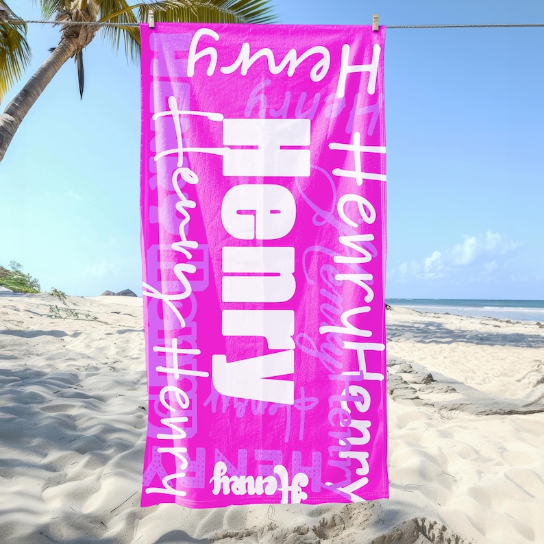 Bright pink personalized beach towel with the name Henry in different fonts, hanging on a line at the beach with palm trees and sand. Custom name towel, perfect for pool, swim, or summer trips for all ages.