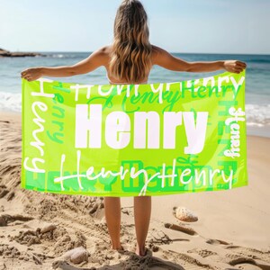 Person stands on a sandy beach holding a bright green personalized beach towel with the name Henry in bold and script fonts. Custom name towel, perfect for pool, swim, or summer trips, and a thoughtful gift for any age.