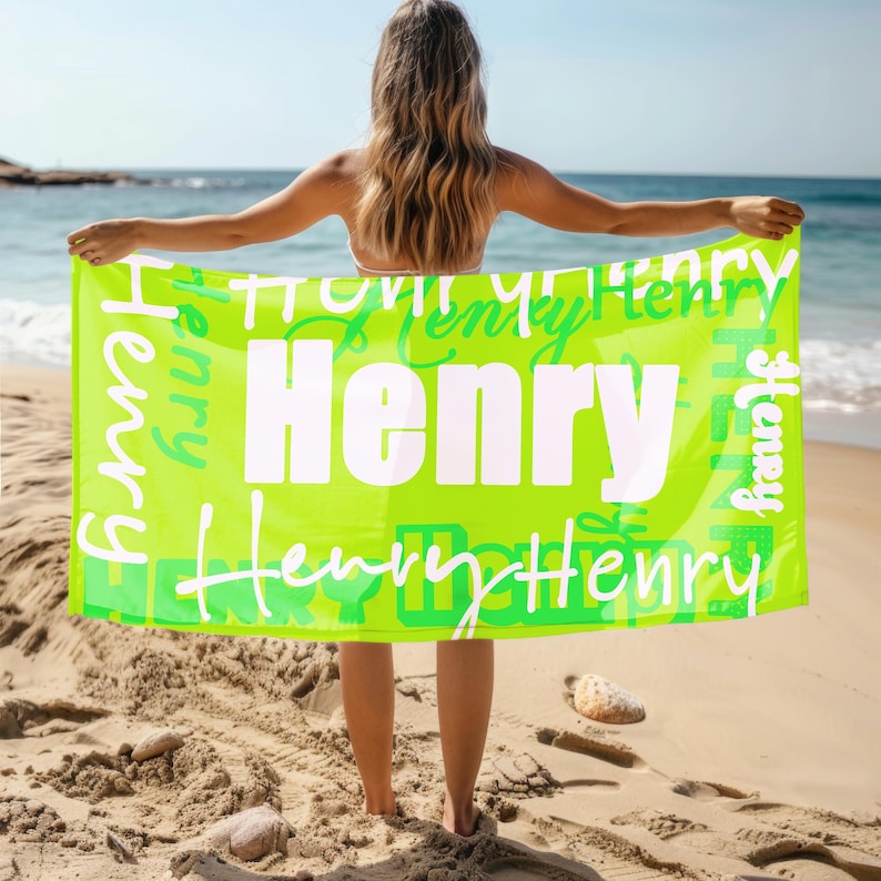 Person stands on a sandy beach holding a bright green personalized beach towel with the name Henry in bold and script fonts. Custom name towel, perfect for pool, swim, or summer trips, and a thoughtful gift for any age.
