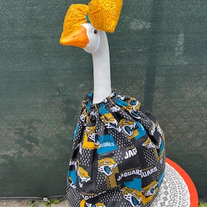 May include: A white goose figurine wearing a yellow fabric bow with a swirling pattern on its head and a gathered skirt featuring a black, teal, and gold Jacksonville Jaguars football team logo print with the text Jaguars visible throughout.