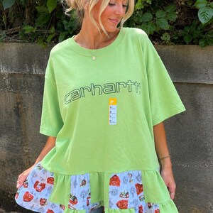May include: A lime green short-sleeve t-shirt featuring the word carhartt printed in dark green across the chest. The bottom hem is accented with a ruffled light blue fabric panel decorated with a repeating pattern of red barns and tractors.