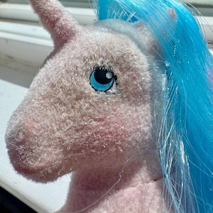Peut inclure: Une peluche licorne avec un corps rose pâle duveteux et une corne unique sur le front. La licorne présente une crinière en cheveux synthétiques bleu vif et de grands yeux ronds avec des iris bleus et des pupilles noires, vue de profil.