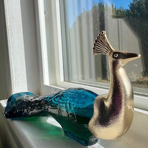 May include: A decorative peacock figurine featuring a metallic gold head and neck with a detailed crest, attached to a translucent teal blue glass body. The glass portion displays a moulded feather pattern across the back and tail section.