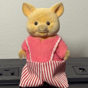 May include: A small, plush pig figurine with a light yellow body and a pink outfit. The outfit includes a pink top with white straps and red and white striped pants. The pig has small black eyes and a smiling expression.