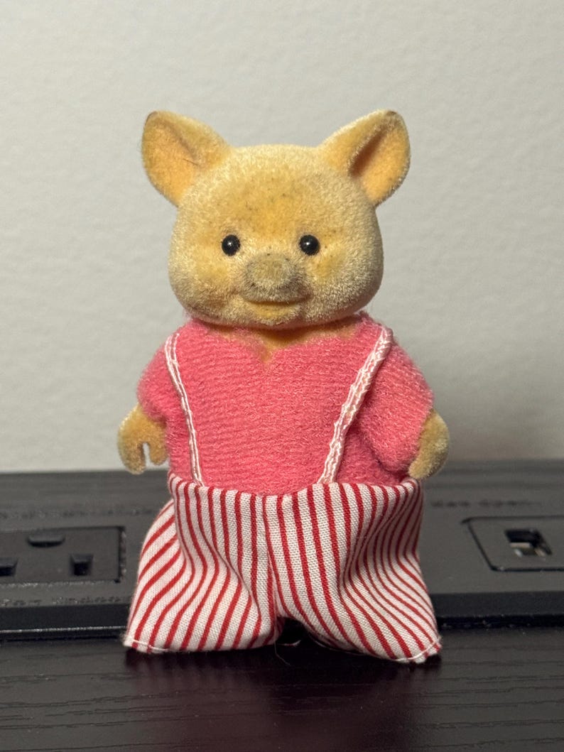 May include: A small, plush pig figurine with a light yellow body and a pink outfit. The outfit includes a pink top with white straps and red and white striped pants. The pig has small black eyes and a smiling expression.